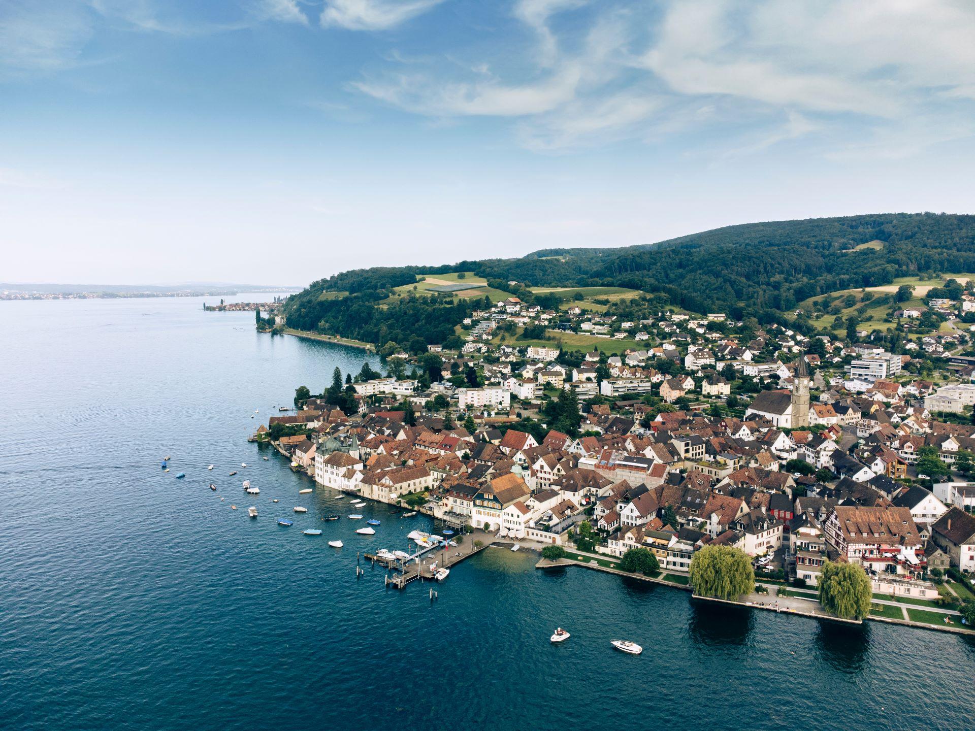 Luftaufnahme eines idyllischen Bodensee-Ortes – Malerische Stadt mit roten Dächern am Bodensee, umgeben von Hügeln und klarem Wasser, mit Booten am Ufer.