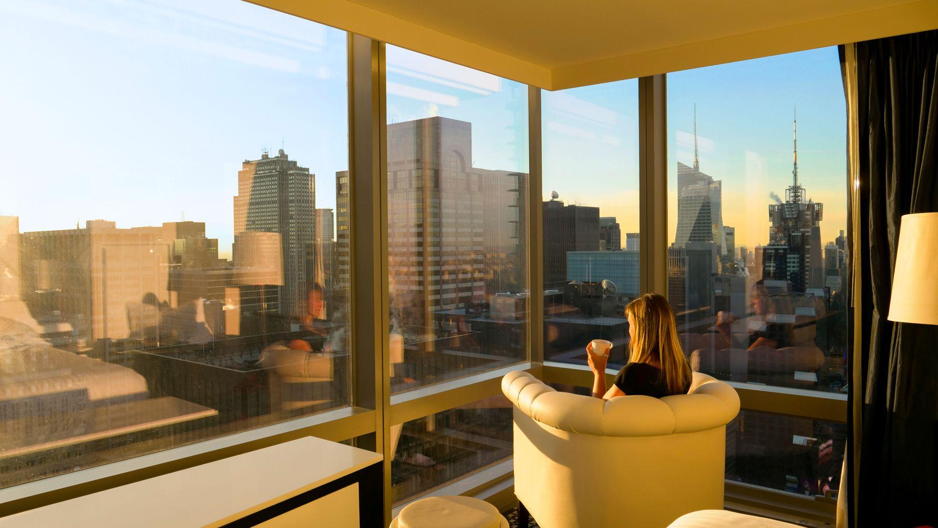 Frau sitzt mit einer Tasse Kaffee in einem hellen Designhotel und blickt auf die moderne Skyline einer Großstadt – ein ruhiger Moment auf einer hochwertigen high class escort Städtereise.